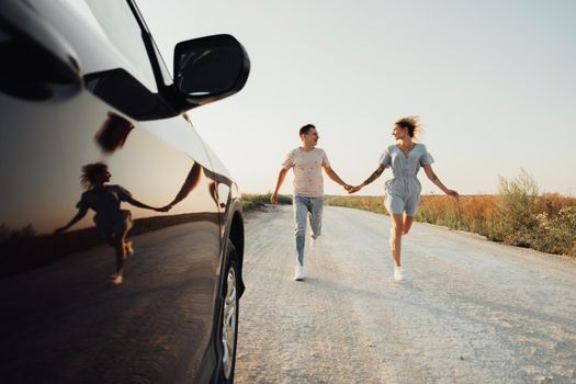 Happy Man and Cheerful Woman Holding by Hands Running Near Car, Young Couple Enjoying Road Trip at Sunset