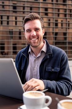 smiling young businessman working with his computer sitting on the terrace of a coffee shop in the financial district, concept of entrepreneurship and business