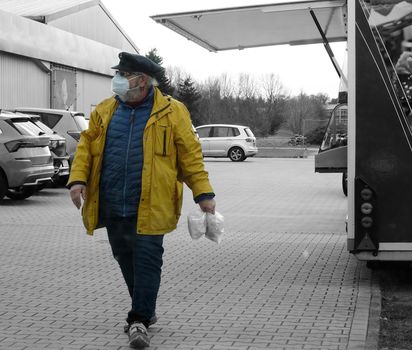 Man with a cap and yellow raincoat goes shopping in times of infection risk. He wears a mask.The colours on this picture are reduced.