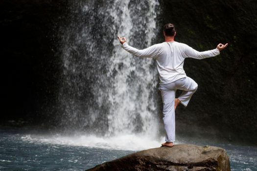 Man standing in meditation yoga on rock at waterfall in tropical Bali