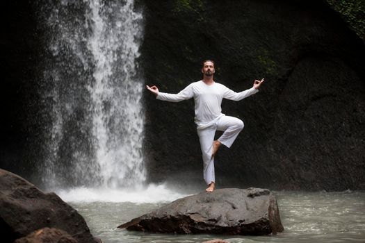 Man standing in meditation yoga on rock at waterfall in tropical Bali