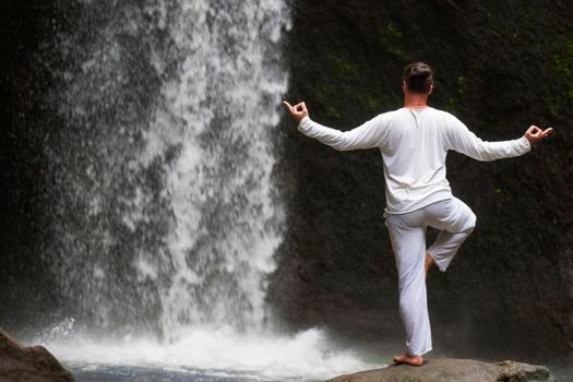 Man standing in meditation yoga on rock at waterfall in tropical Bali