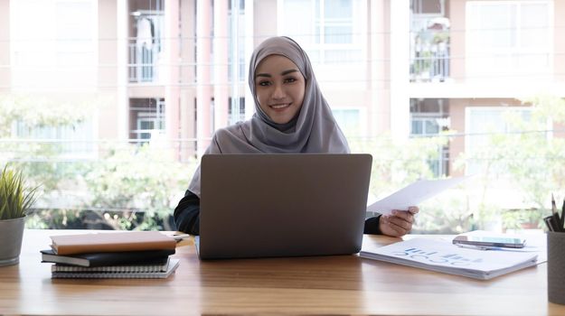 Asian Muslim hijab woman using laptop computer at work smiling at camera.