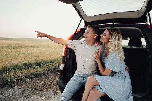 Cheerful Woman and Man Sitting in Trunk of SUV Car, Happy Couple Enjoying Road Trip at Sunset