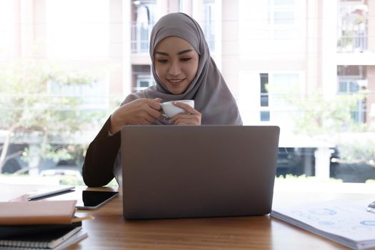 Asian Muslim hijab woman holding coffee cup looking at computer smiling happily.