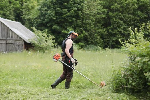 Worker man mowing tall grass with petrol lawn trimmer in the garden or backyard. Process of lawn trimming