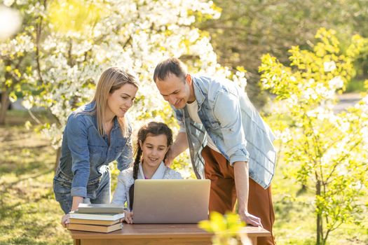 Smiling young Caucasian family with small daughter look at laptop screen watching funny video online. Happy mom and dad have fun using computer with little girl child outdoor. Technology concept