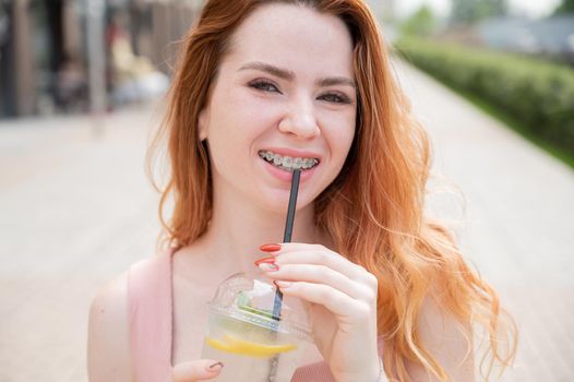 Young beautiful red-haired woman with braces drinks cooling lemonade outdoors in summer. Portrait of a smiling girl with freckles