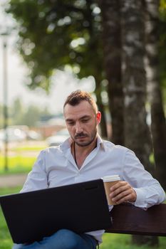 Concentrated man works in the park with a laptop, drinks coffee. A young man on a background of green trees, a hot sunny summer day. Warm soft light, close-up.