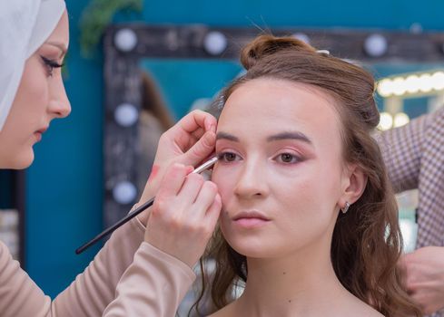 Make-up artist with a brush paints the eyelid close-up of a girl in the salon. Female master makes makeup to a young woman. Business concept - beauty salon, facial skin care, cosmetology.