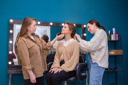 Two masters neatly style the hair and make up the girl in the salon. Make-up artist and hairdresser create an image of a young woman. Business concept - beauty salon, facial skin and hair care.