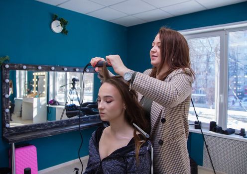 A young woman master with a smile twists a strand of the client's hair on tongs. The hairdresser makes a hairstyle for a young woman. Barber shop, business concept. Beauty salon, hair care.