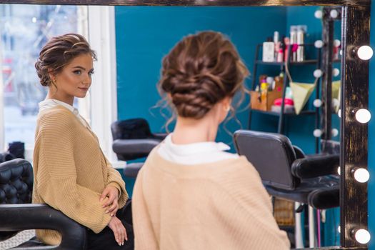 A girl with fashionable makeup and hairstyle looks in a large mirror. A new look for a young woman. Business concept - beauty salon, facial skin and hair care.