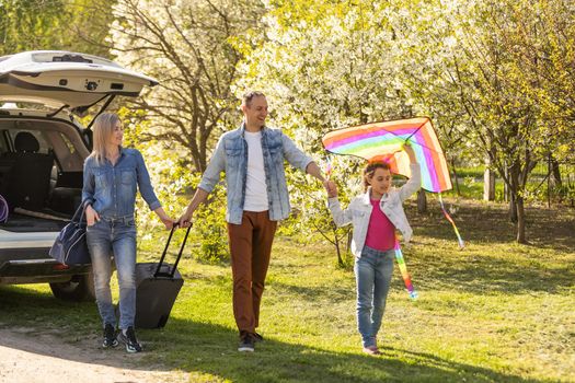 Happy family standing together near a car with open trunk enjoying view of rural landscape nature. Parents and their kid leaning on vehicle luggage compartment. Weekend travel and holidays concept