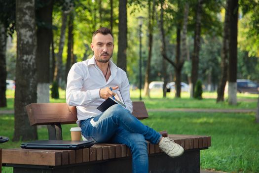 A serious man sits on a bench, makes plans, writes in a notebook. A young man on a background of green trees, a hot sunny summer day. Warm soft light, close-up.