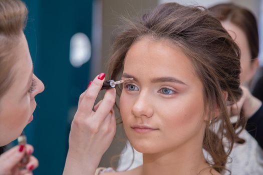 Make-up artist with a brush paints the eyebrows close-up of a girl. Female master makes makeup to a young woman. Business concept - beauty salon, facial skin care, cosmetology.