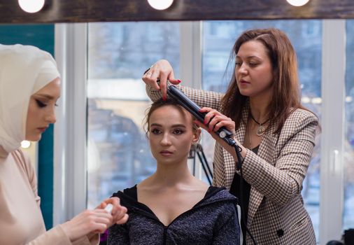 Two women professionally curl hair and make up a girl in a salon. Make-up artist and hairdresser create an image of a young woman. Business concept - beauty salon, facial skin and hair care.