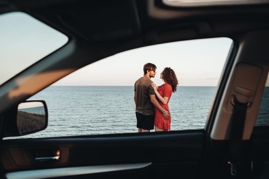 View Through Window of the Car Young Couple Standing on Background of Sea, Man Hugging Curly Woman in Red Dress at Sunset