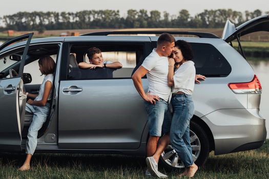 Caucasian Man and Woman Hugging Near the Minivan Car, Happy Four Members Family Enjoying Road Trip, Making Stop by River