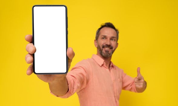 Mature grey haired man showing huge smartphone in hand happy smiling looking at device wearing peach shirt isolated on yellow. Mature fit man with phone app advertisement. Selective focus on phone.