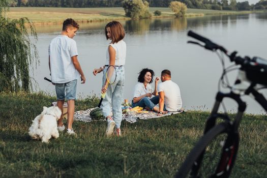 Middle Aged Man and Woman Having Picnic While Their Two Teenage Children, Son and Daughter Playing with Pet Dog, Happy Four Members Family Enjoying Weekend Outdoors by Lake