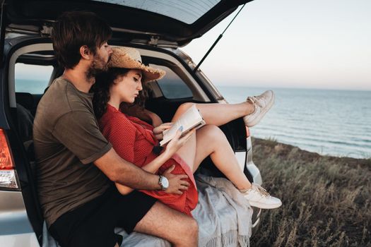 Man and Woman Sitting Inside Opened Trunk of SUV Car, Young Couple Enjoying Road Trip Along Sea at Sunset