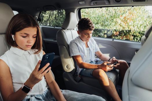Young Caucasian Boy and His Teenage Sister Focused on Smartphones While Sitting in Minivan, Children Playing Games on Weekend Family Road Trip by Car