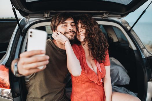 Happy Caucasian Couple Man and Curly Brunette Woman Making Selfie on Smartphone While Sitting in Trunk of SUV Car
