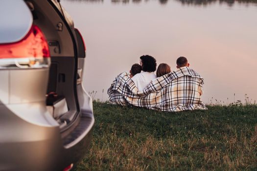 Four Members Family Sitting on Grass by the Lake at Sunset, Mother and Father with Two Children Enjoying Weekend Road Trip