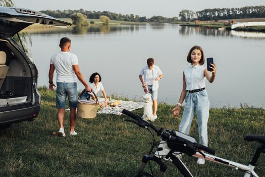 Happy Four Members Family Enjoying Weekend Road Trip by Minivan Car, Teenage Daughter Making Selfie While Her Brother Playing with Pet Dog by Lake