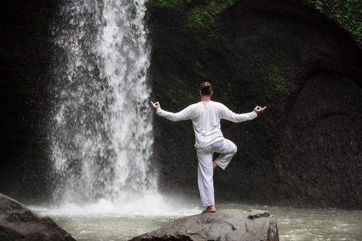 Man standing in meditation yoga on rock at waterfall in tropical Bali