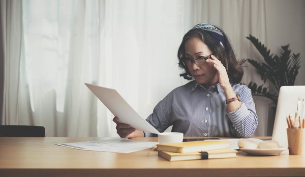 Seriously senior woman tworking at home, looking at laptop and waving paper correspondence, sitting at home, free space..