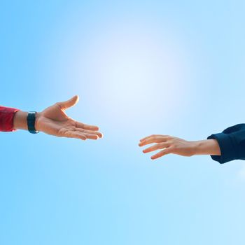 Shot of two unidentifiable hikers reaching for each others hands against a clear blue sky.