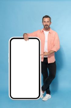 Middle aged grey haired man leaned on huge, giant smartphone pointing on white screen happy smiling on camera wearing peach shirt isolated on blue background. Mature fit man with smartphone.