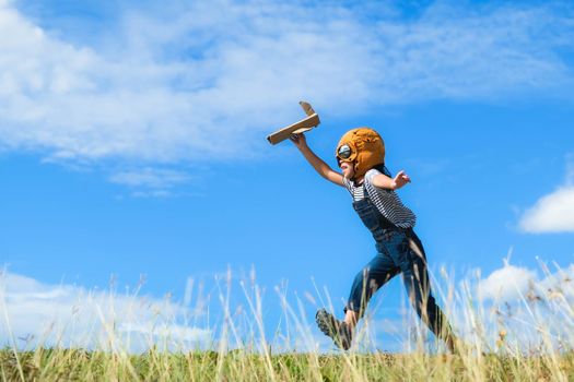 Cute little girl running through the meadow on a sunny day with a toy plane in hand. Happy kid playing with cardboard plane against blue summer sky background. Childhood dream imagination concept.