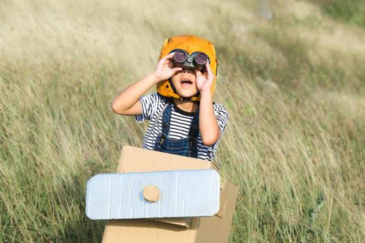 Cute dreamer little girl playing with cardboard planes in the meadow on a sunny day. Happy kid playing with cardboard plane against blue summer sky background. Childhood dream imagination concept.