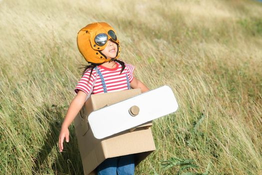 Cute dreamer little girl playing with cardboard planes in the meadow on a sunny day. Happy kid playing with cardboard plane against blue summer sky background. Childhood dream imagination concept.