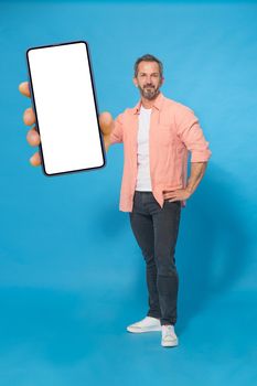 Grey haired middle aged man gesturing hello with one hand up happy smiling on camera wearing peach color shirt and grey jeans isolated on blue background. Mature fit man, healthy lifestyle concept.