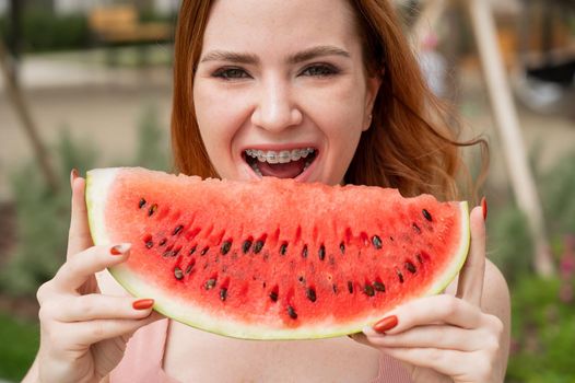 Beautiful red-haired woman smiling with braces and about to eat a slice of watermelon outdoors in summer.