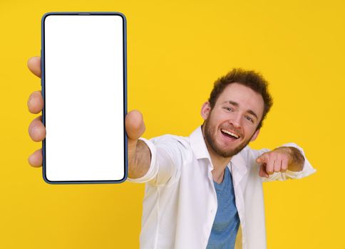 Happy handsome young guy with smartphone in hand showing a white empty screen, cellphone display mock up isolated on yellow background. Product placement mobile app advertisement.