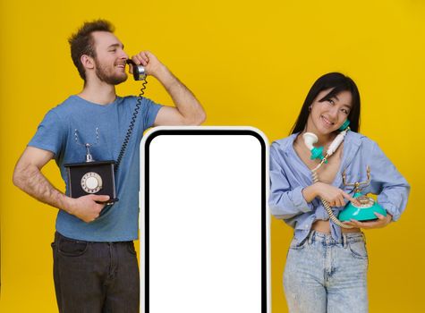 Handsome guy and asian girl talking on vintage phones leaned on huge smartphone or digital tablet with blank screen, happy smiling isolated on yellow background. Mock up, product placement.