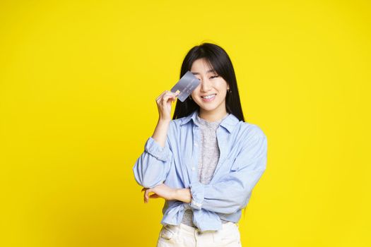 Charming asian girl holding credit, debit card touching her eye. Asian girl in blue shirt holding mockup credit card isolated on yellow background. Shopping concept. Banking concept.