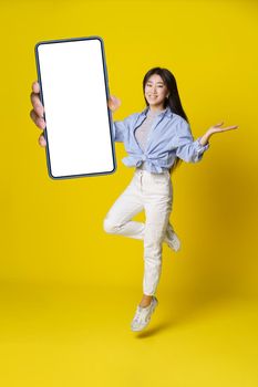 Asian girl in jump holding smartphone with black screen in hand in casual outfit, mobile app advertisement isolated on yellow background. Full length portrait of joyful asian girl. Product placement.