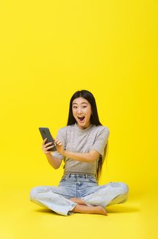 Excited asian young girl sitting on the floor holding smartphone texting or shopping online or win gaming isolated on yellow background. Product placement. Mobile App Advertisement. Copy space.