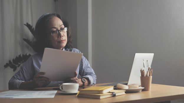 Seriously senior woman tworking at home, looking at laptop and waving paper correspondence, sitting at home, free space..