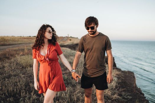 Cheerful Couple Walking Along Sea, Man and Curly Woman in Red Dress Holding by Hands Enjoying Their Vacation