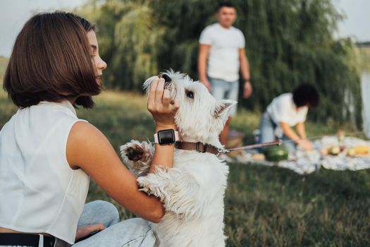 Close Up Teenage Girl Playing with West Highland White Terrier Dog on Background of Her Family Having Picnic Outdoors