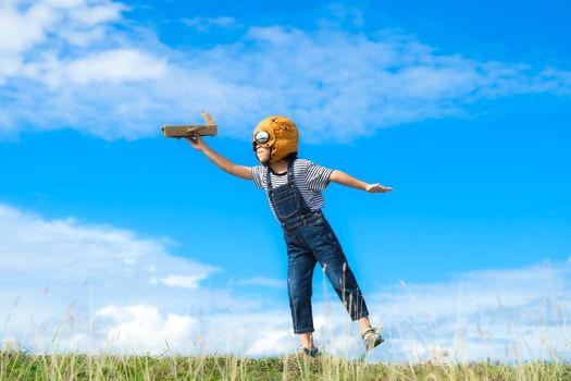 Cute little girl running through the meadow on a sunny day with a toy plane in hand. Happy kid playing with cardboard plane against blue summer sky background. Childhood dream imagination concept.