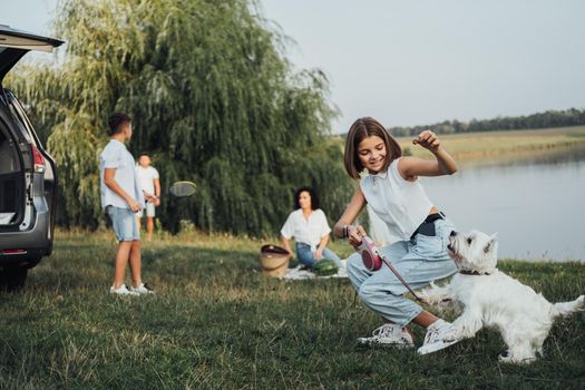 Teenage Girl Playing with West Highland White Terrier Dog on Background of Her Family Having Fun Time Outdoors on Road Trip by Minivan Car