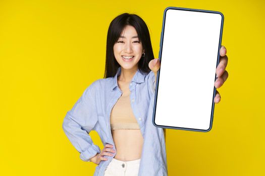 Asian girl holding smartphone showing white screen, and excited smile on camera wearing blue shirt isolated on yellow background. Great offer mobile app advertisement. Product placement.